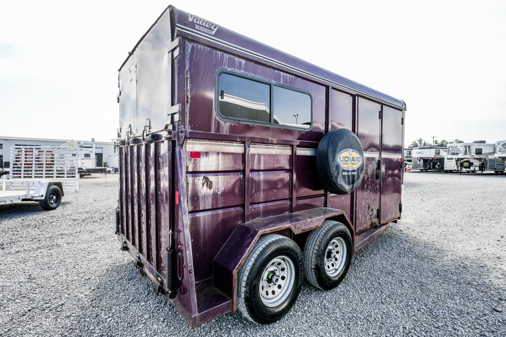 Maroon double-axle horse trailer with a side spare tire on a gravel lot, reflections in the window.