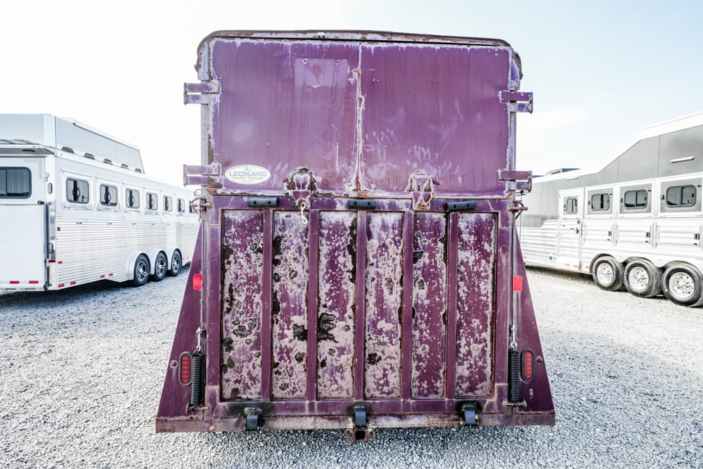 Rear view of a weathered purple trailer with peeling paint and rust, parked on a gravel lot among other horse trailers.