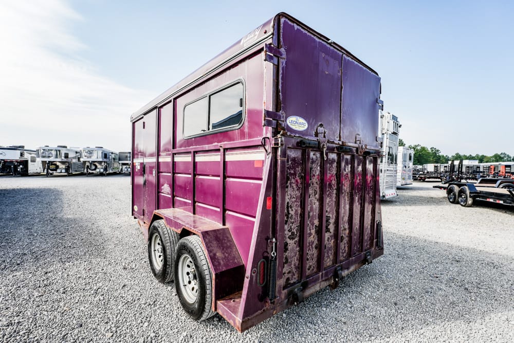 Rear view of a purple two-axle livestock trailer on a gravel lot, with other trailers in the background.