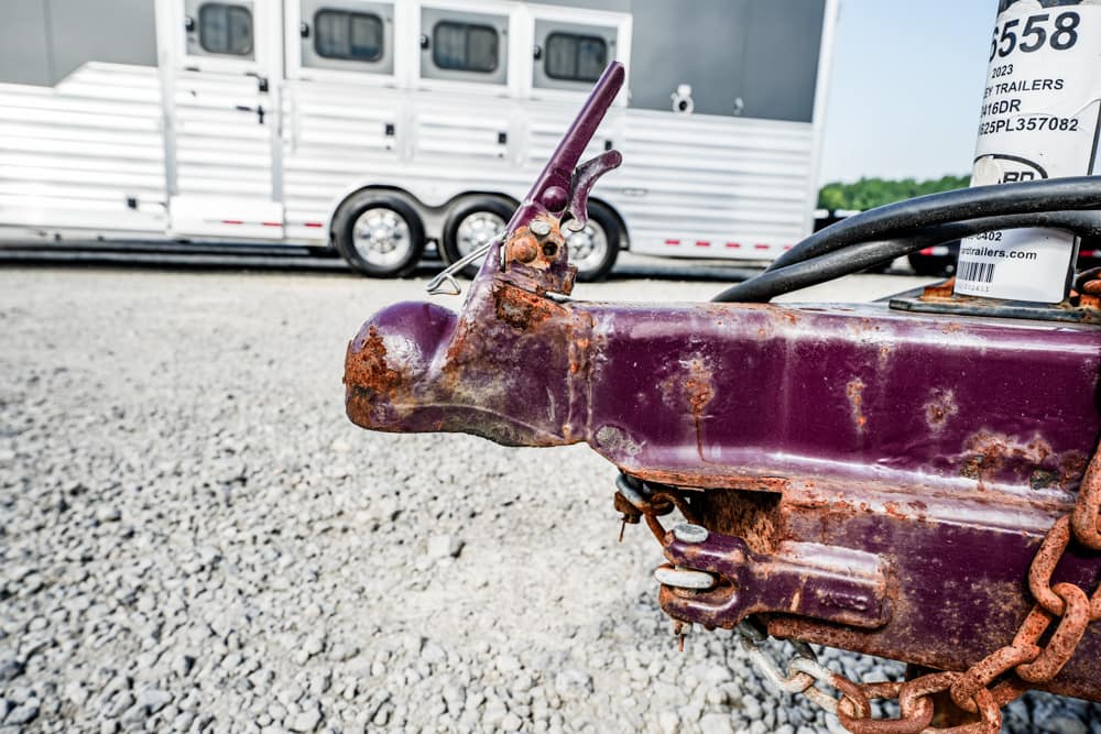 Close-up of a rusty purple trailer hitch with chains on a gravel lot; a horse trailer is blurred in the background.