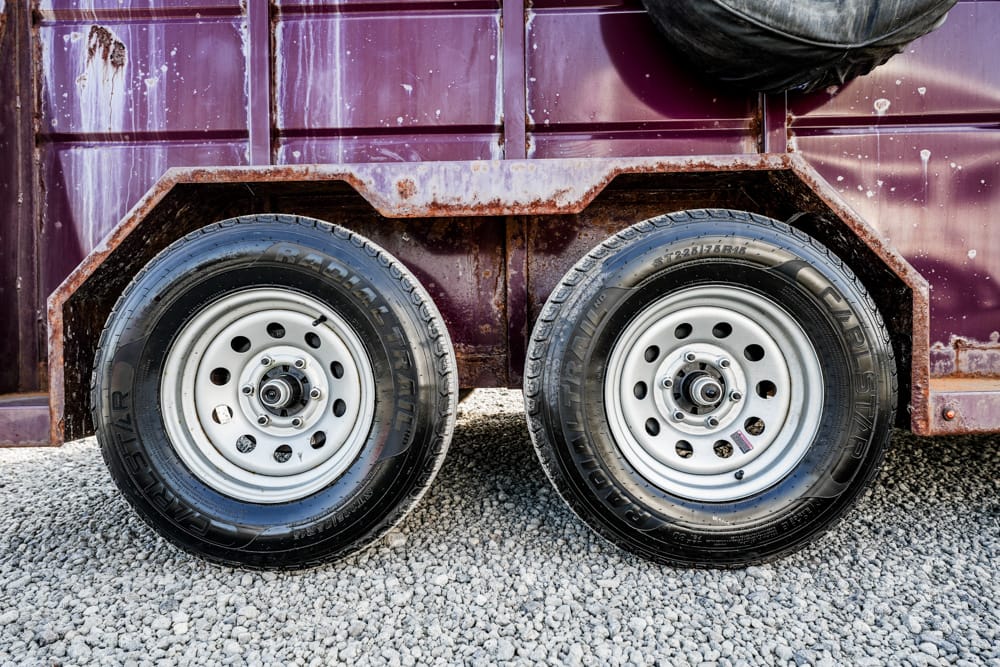 Dual-axle trailer with two black tires and silver rims on a gravel surface, a rusted purple metal body in the background, with a spare tire visible on top