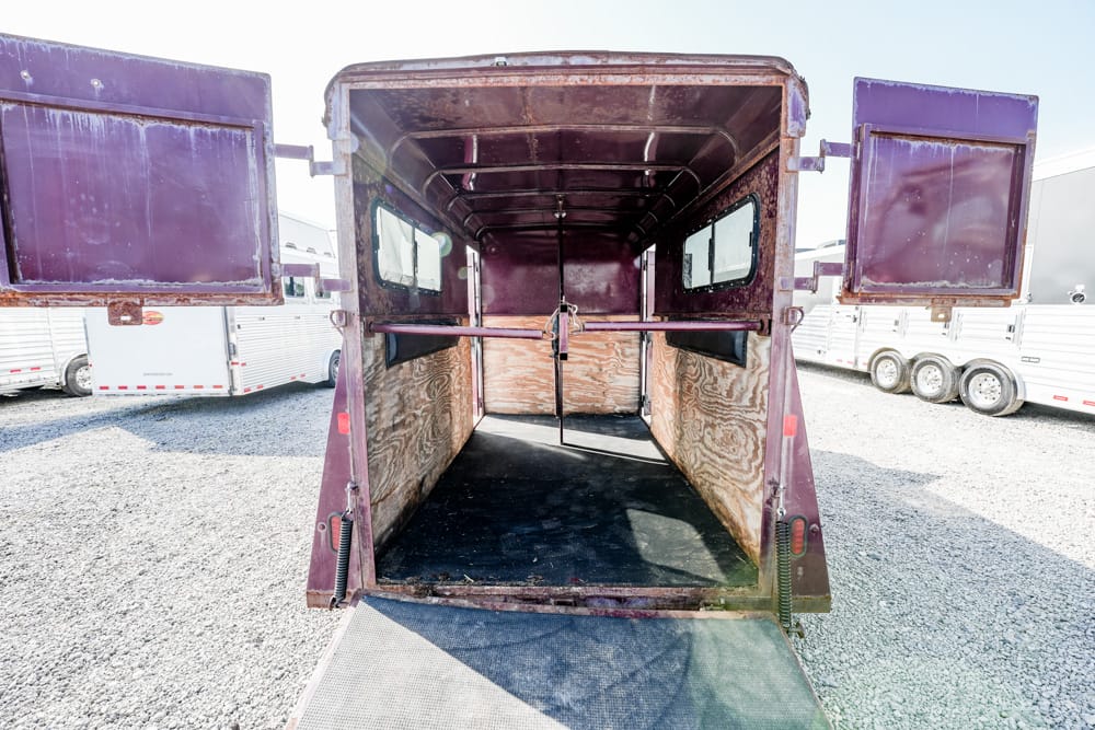 Open maroon horse trailer interior with a center divider and wooden walls, ramp down on a gravel lot