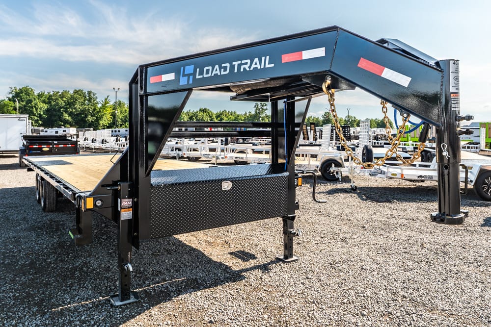 Load Trail gooseneck flatbed trailer with wood deck, diamond-plate toolbox, and safety chains on a gravel lot.