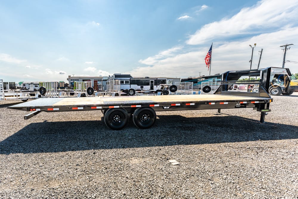 Flatbed trailer with dual axles on a gravel lot, surrounded by other trailers and an American flag in the background.