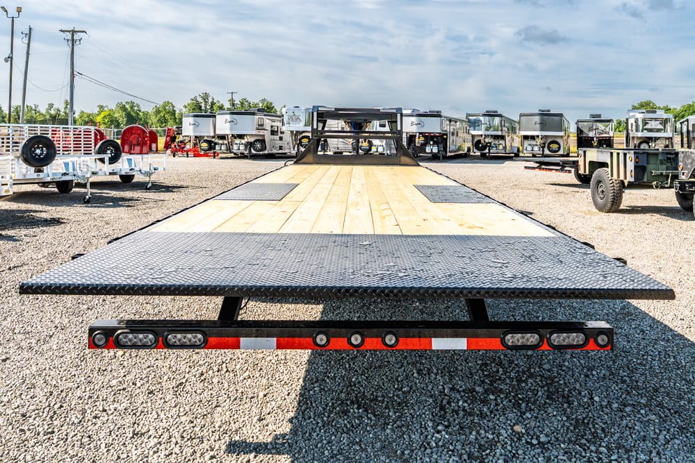 Low-angle view of a flatbed trailer with wooden deck and diamond-plate edges in a gravel truck yard, with trailers in the background.