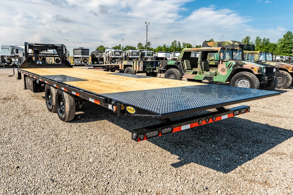 Flatbed trailer with a diamond-plate ramp on a gravel lot, military trucks visible in the background