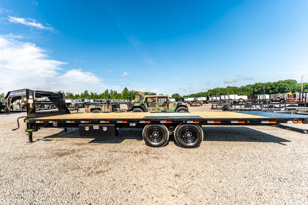 Black flatbed trailer with dual rear axles on a gravel lot, with other trailers and a camouflaged military vehicle in the background.