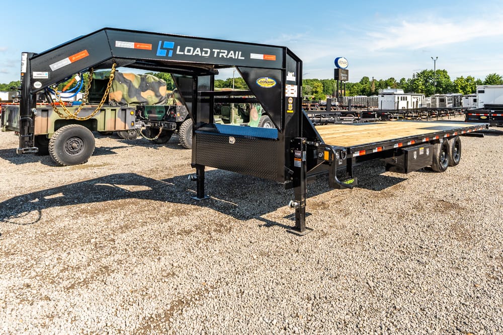 Black Load Trail gooseneck flatbed trailer on a gravel lot with a long wood/steel deck and dual rear axles, brand visible on frame
