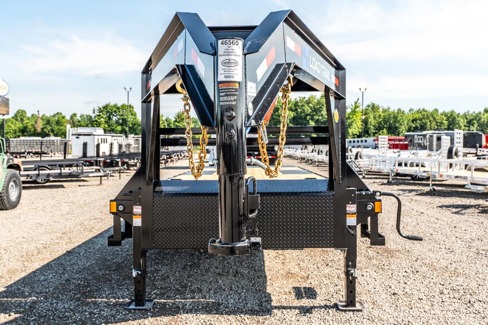 Front view of a black industrial trailer frame with safety chains and a gooseneck hitch, parked on a gravel lot with other trailers in the background, sunny day.