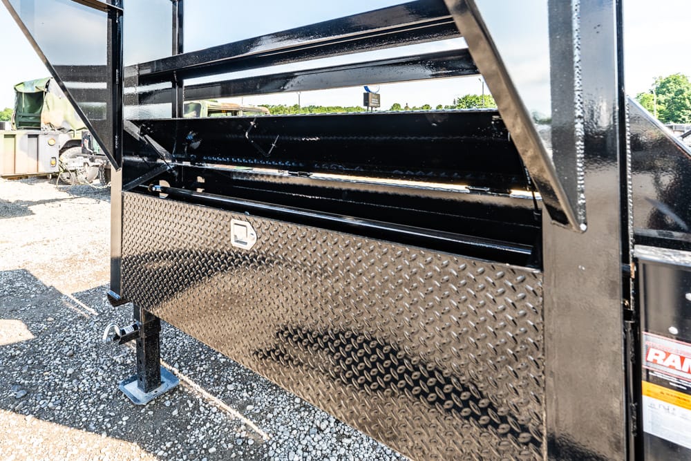 Close-up of a black diamond-plate utility trailer side, showing a latch, support leg, and reflective metal surface on gravel ground.