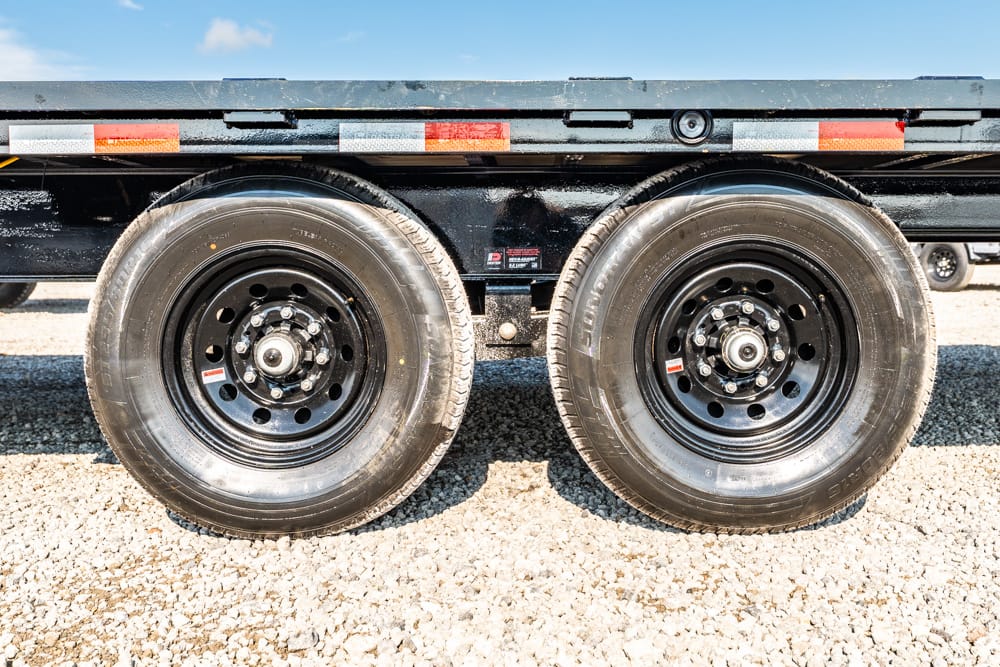 Dual heavy-duty trailer wheels on a gravel surface beneath a flatbed trailer, showing lug nuts and rims under clear blue sky.