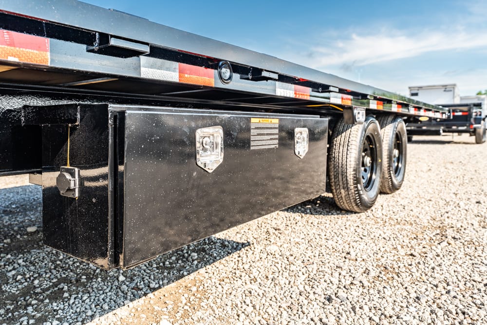 Black metal storage box mounted under a flatbed trailer, with dual rear wheels and a gravel lot in the background.
