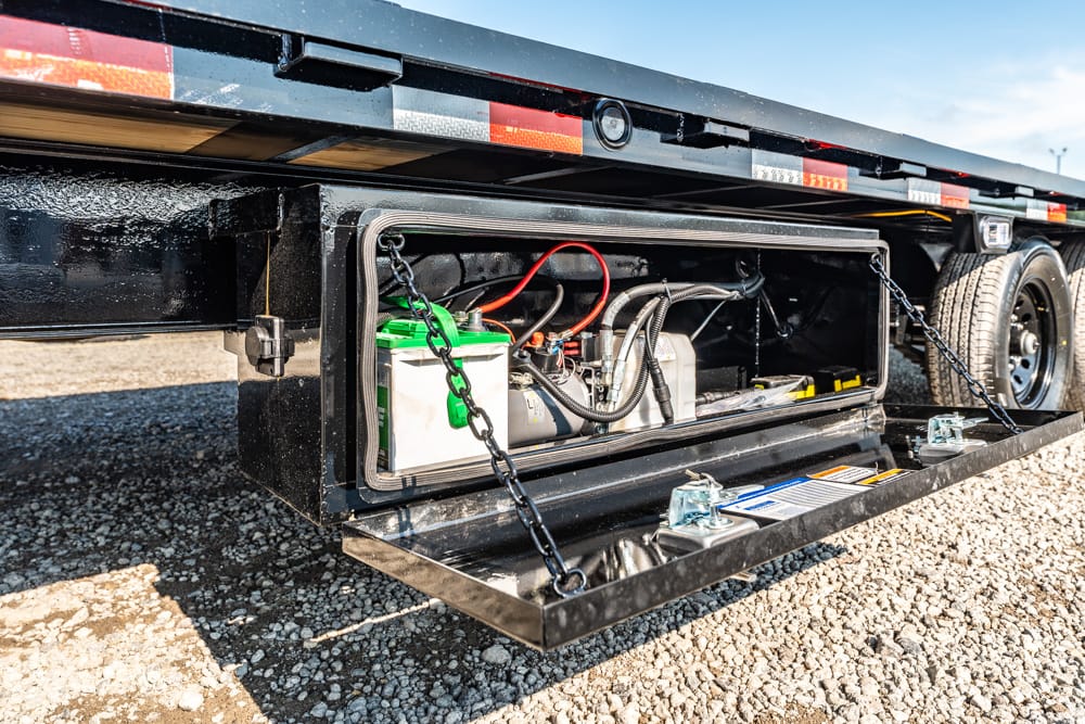 Open storage compartment of a flatbed trailer with a battery, hoses, and cables inside.