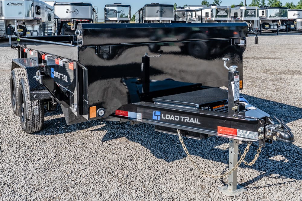 Black Load Trail dump trailer on a gravel lot with a front hitch and safety chains in the foreground, cargo bed raised.