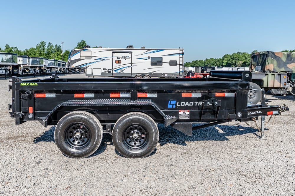 Black dual-axle Load Trail dump trailer on a gravel lot, with RVs and a camo truck in the background