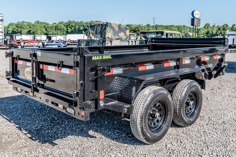 Black dual-axle dump trailer with high side walls in a gravel lot; 'MAX RAIL' and 'LOAD TRAIL' branding visible.
