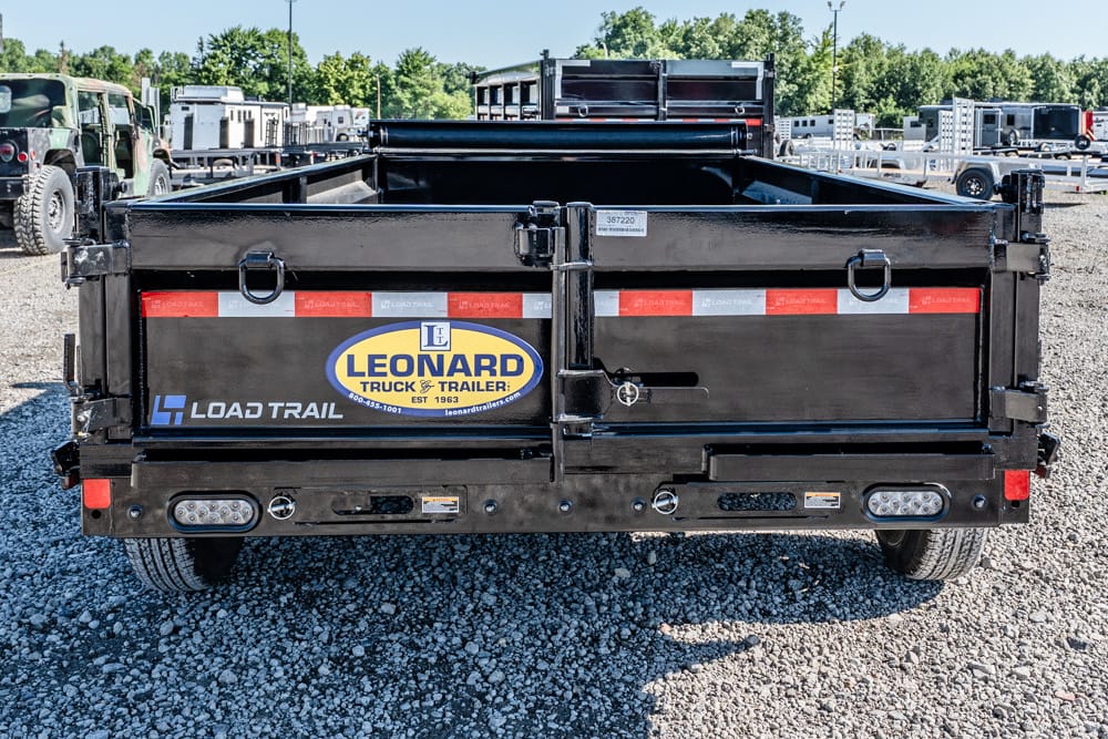 Black Load Trail dump trailer back view with Leonard Truck & Trailer logo in a gravel lot. Full-width tailgate closed, red reflective strip along top, headlights and lights at bottom edge visible.