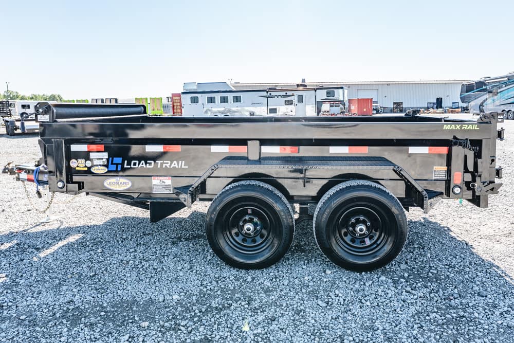 Black Load Trail dump trailer with dual axles on a gravel lot, rear gate visible, with RVs and a warehouse in the background.