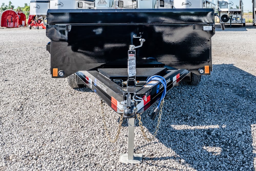 Front end of a black utility trailer on a gravel lot, showing the hitch, safety chains, and a wheel jack nearby.
