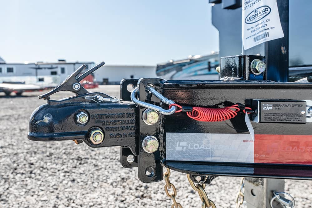 Close-up of a trailer hitch with a safety latch and red coiled cord on a gravel lot, outdoors in bright sunlight.