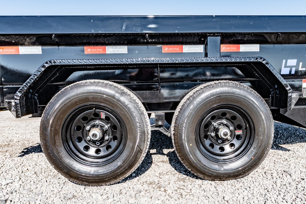 Close-up of a tandem-axle dump trailer showing two black steel wheels on a gravel surface under a black metal frame.