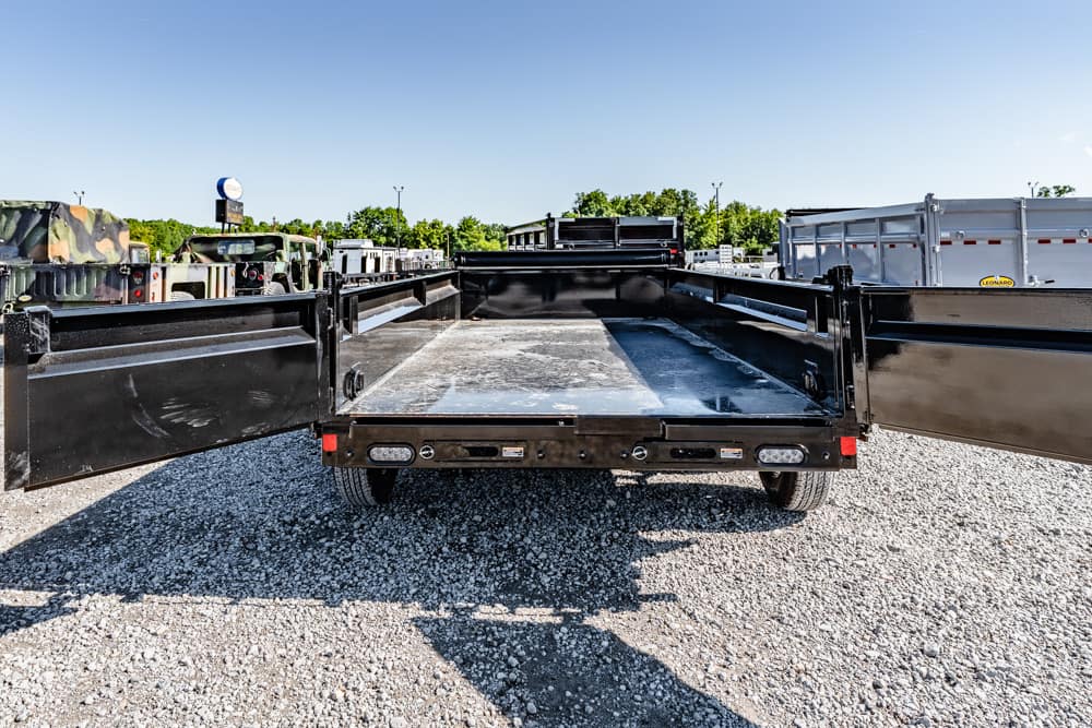 Black flatbed trailer with open side gates, empty bed, parked on gravel lot among other trucks and trailers.