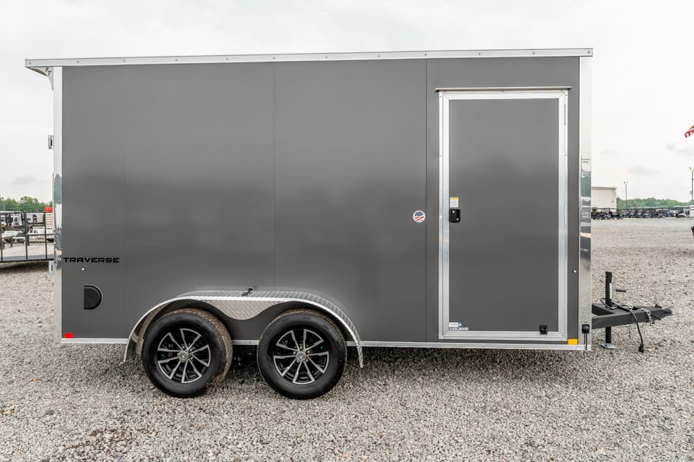 Gray enclosed cargo trailer with dual axles and diamond-plate fenders on a gravel lot, side door closed