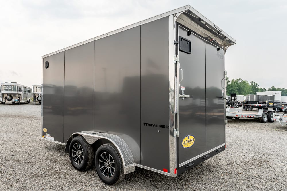 Gray two‑axle enclosed utility trailer on a gravel lot with diamond‑plate fenders and rear doors closed, brand name 'Traverse' visible on the side