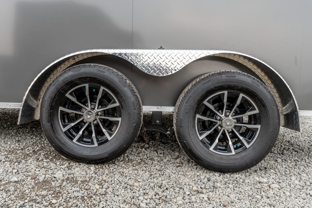 Dual-axle trailer wheels with diamond-plate fenders on a gravel surface.