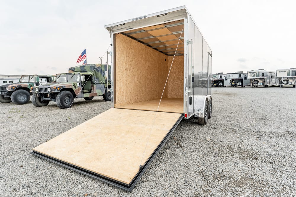 Open cargo trailer with plywood interior and ramp lowered on a gravel lot, military vehicles in the background.
