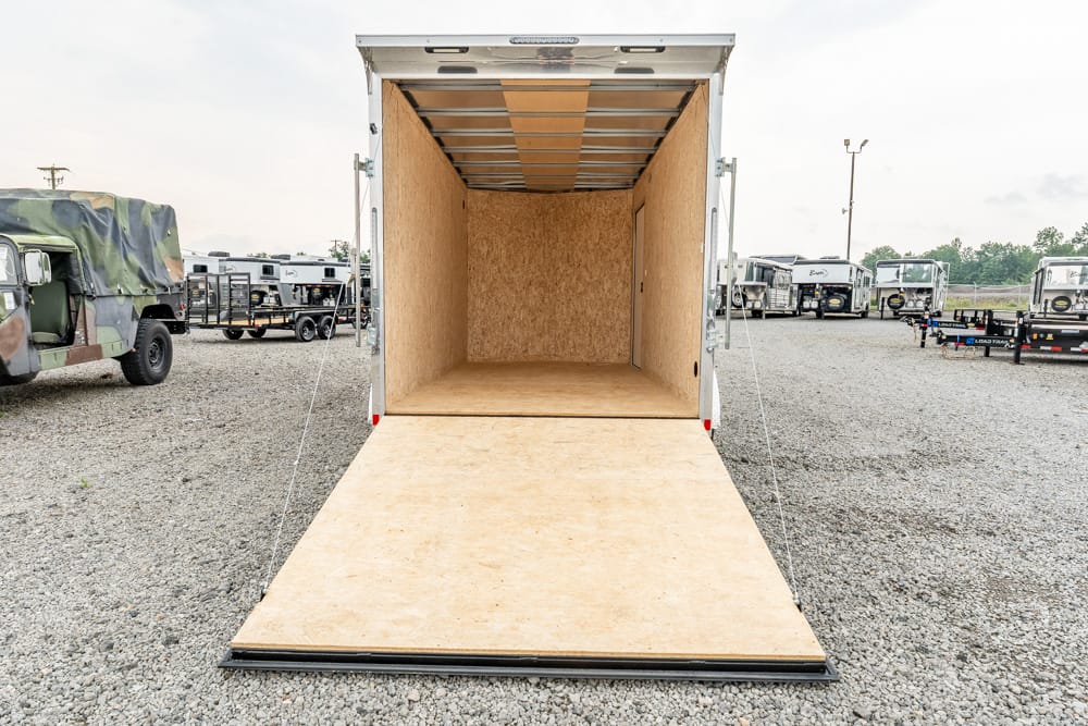 Open cargo trailer with ramp down, empty plywood interior, parked on a gravel lot.