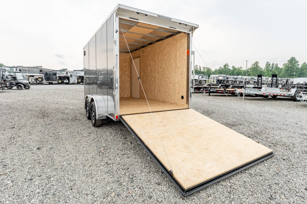 Silver enclosed cargo trailer with the ramp lowered to ground in a gravel lot, other trailers in the background.