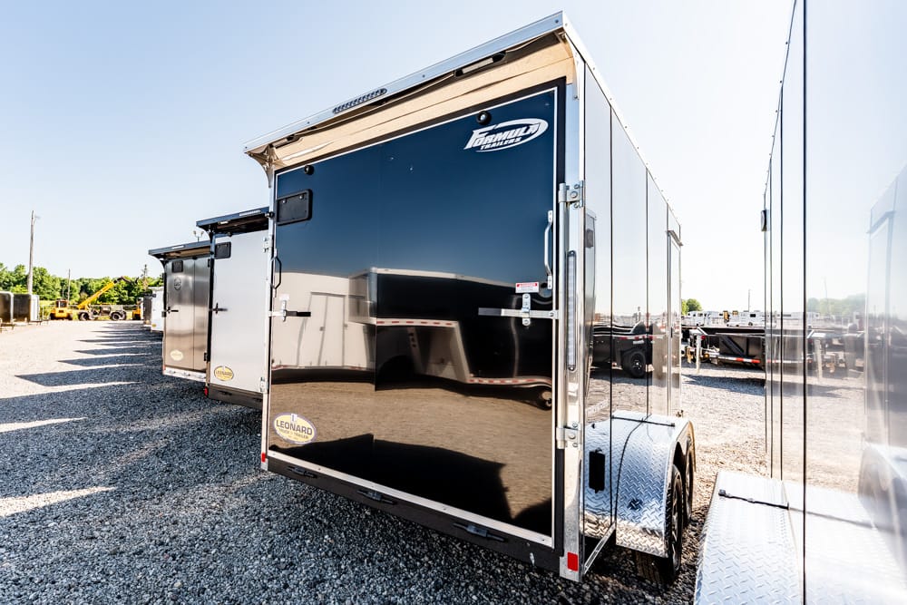 Shiny black enclosed cargo trailer with dual axles parked on a gravel lot, its surface reflecting surrounding trailers and sunlight.] ,