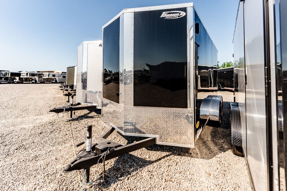Row of enclosed cargo trailers on a gravel lot; the foreground trailer has a black reflective front with diamond-plate trim and a hitch in view.
