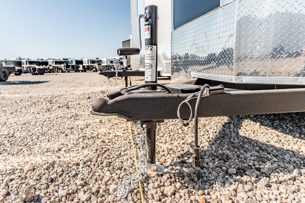 Close-up of a trailer tongue with a hitch and jack on a gravel lot, with a diamond-plate trailer body to the right and other trailers in the background, during daylight.