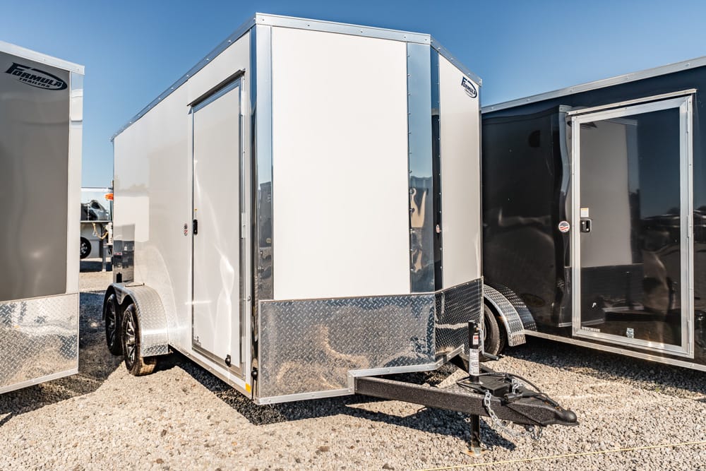 White enclosed cargo trailer with diamond-plate front and tandem axles parked on a gravel lot beside other trailers.