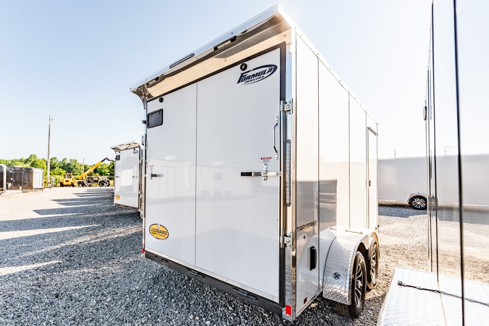 White enclosed cargo trailer with tandem wheels parked on a gravel lot; rear doors closed and a small logo visible on the back door edge.
