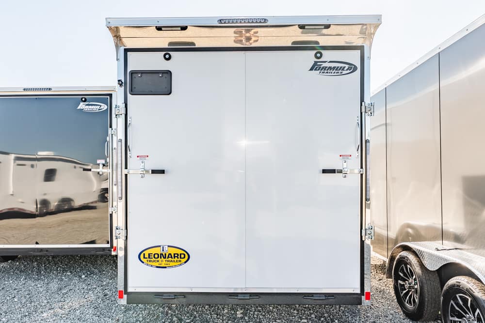 Rear of a white enclosed trailer with two doors and Leonard logo at the bottom left on a gravel lot.