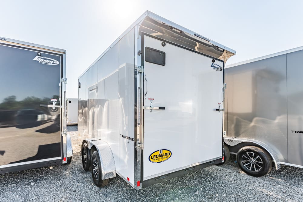 White two‑axle enclosed cargo trailer with diamond‑plate fenders, parked on a gravel lot between other trailers.