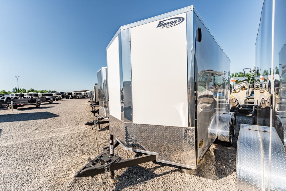 Line of shiny enclosed cargo trailers parked on a gravel lot under a clear blue sky, dealership row stretching into the distance