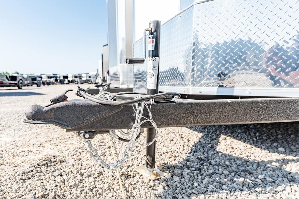 Close-up of a trailer hitch with safety chains and jack on a gravel lot, with a diamond-plate trailer body in the background.