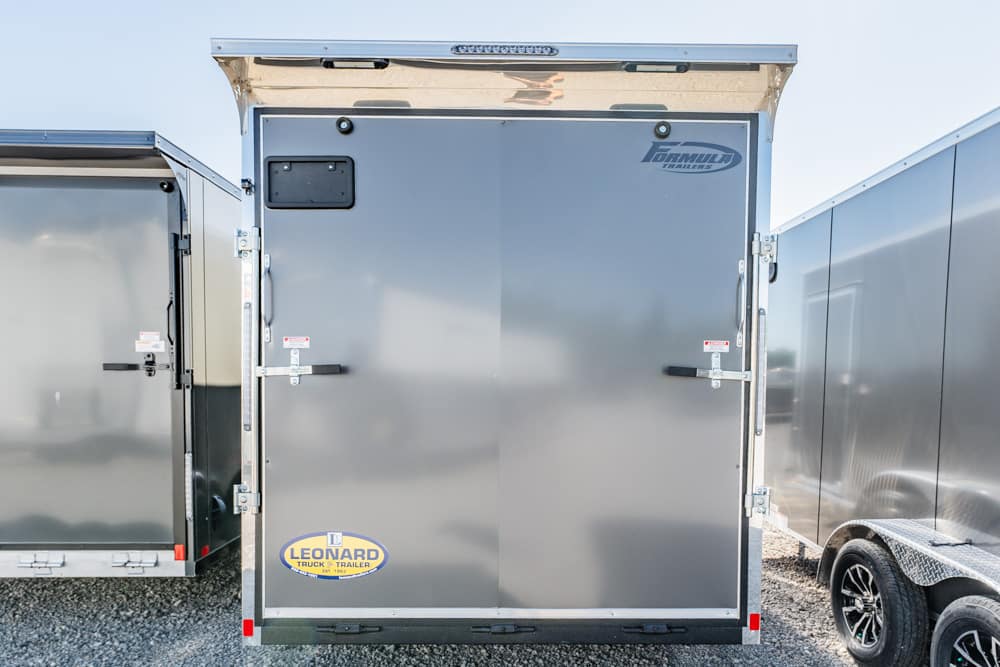 Rear view of a gray enclosed cargo trailer with double doors and visible Leonard trailer logo on a gravel lot.
