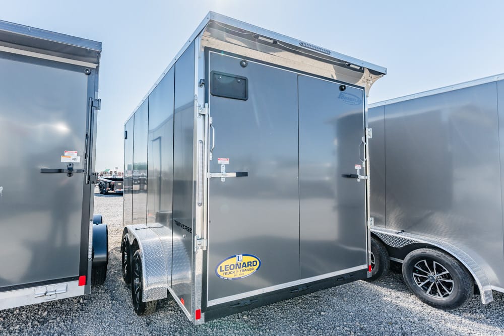Gray enclosed cargo trailer with dual rear doors, tandem wheels, and diamond-plate fenders on a gravel lot.