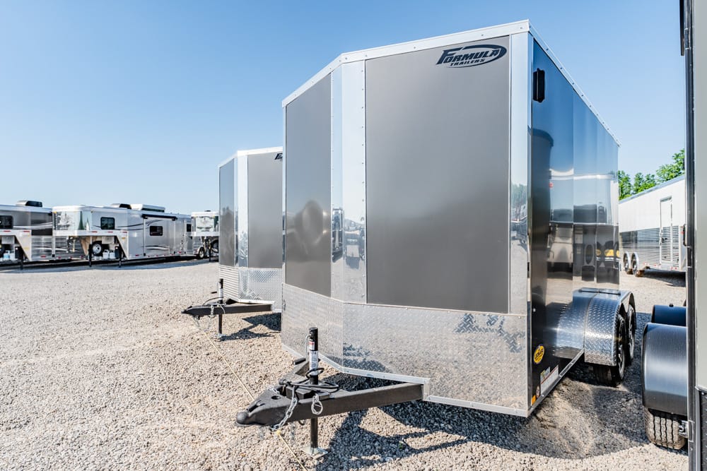 Row of shiny enclosed cargo trailers on a gravel lot, with front tongues visible and diamond-plate trim around the base, under a clear blue sky.
