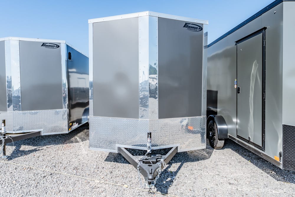 Front view of a gray enclosed cargo trailer with diamond-plate trim, hitched to the hitch on a gravel lot.