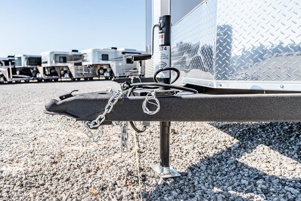 Close-up of a trailer tongue with hitch, safety chains, and a jack on a gravel surface, with other trailers in the background.
