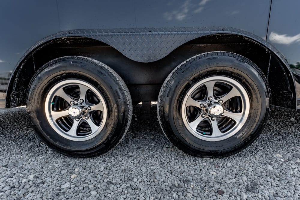 Pair of silver alloy wheels on a black trailer, resting on gravel surface beneath a curved diamond-plate fender.