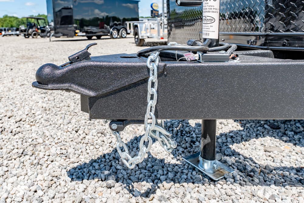 Close-up of a black trailer hitch with a heavy safety chain looped around the tongue, resting on a gravel lot.
