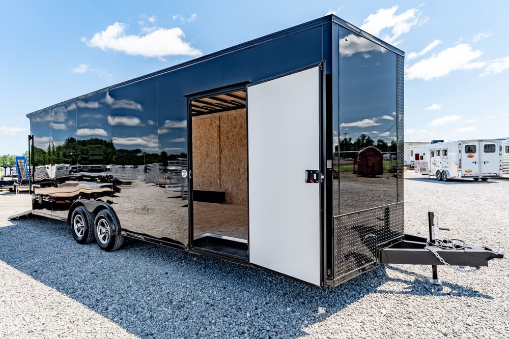 Black enclosed cargo trailer with dual axles, hitch at front, and open side door showing unfinished plywood interior on a gravel lot