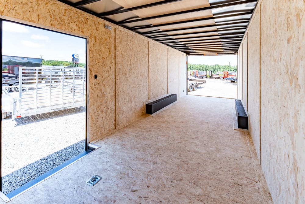 Interior of an empty plywood trailer with benches along both walls and an open rear exit to a yard outside.